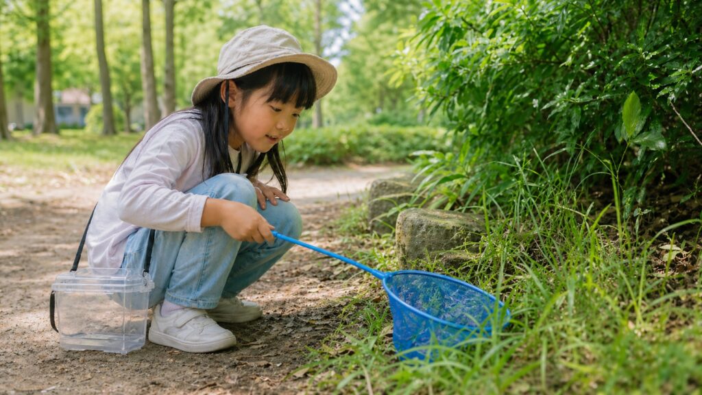 公園で長袖と長ズボン姿の日本人の女の子が、虫取り網を手に地面近くの虫を観察している様子