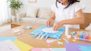 小学生の女の子がリビングのテーブルで色紙やのりを使って工作をしている様子