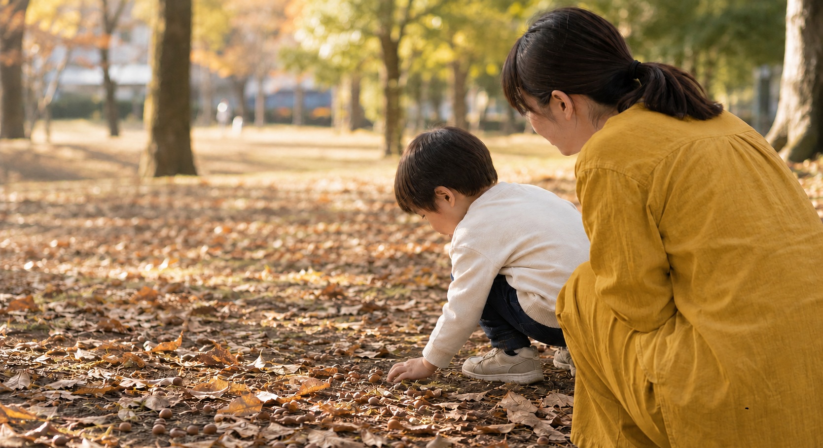秋の公園でどんぐりを見つける3歳くらいの男の子とそれを見守る母親