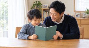 ダイニングでブックカバーをかけた図鑑を楽しそうに読む父親と小学生の男の子